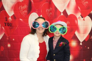 mom and son posing for a picture in front of a heart backgound at the sweetheart dance.